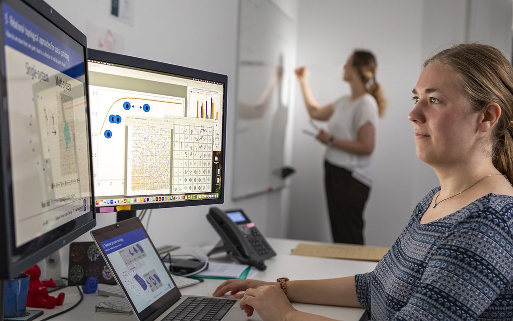 Woman working at a computer with multiple monitors; another person is standing in front of a board in the background.