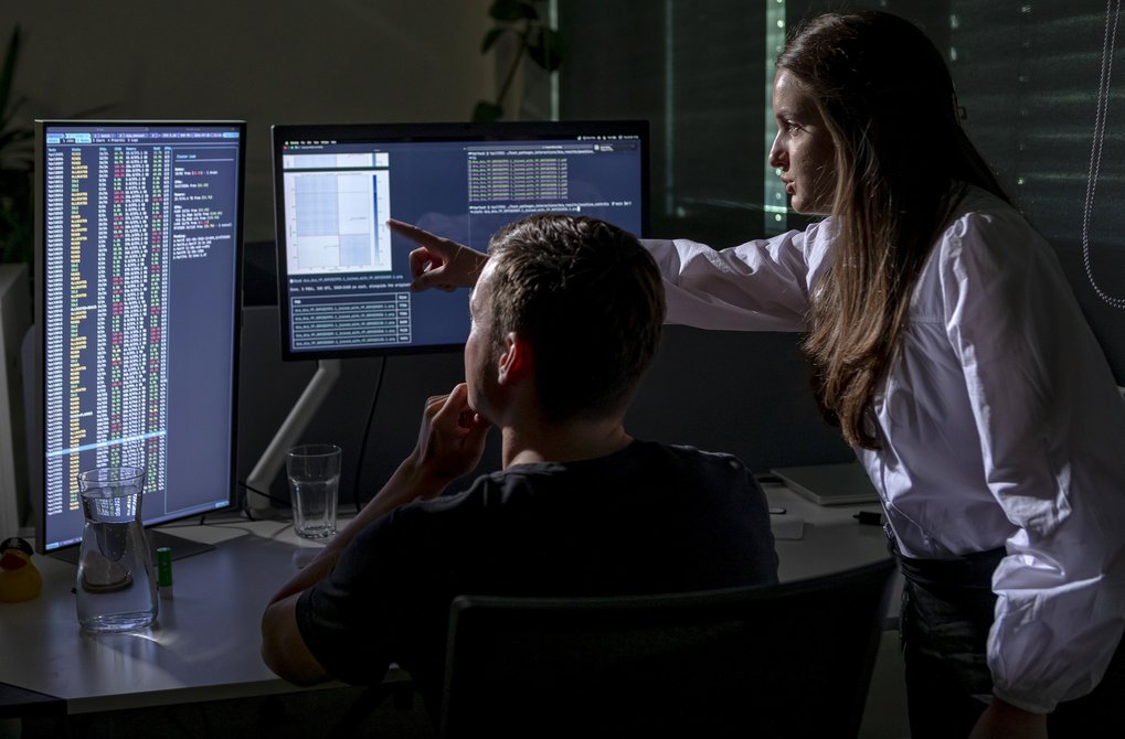 In a low-lit workspace, a female professional directs attention to coding and graphical data on one of two monitors as a male colleague contemplates the information.